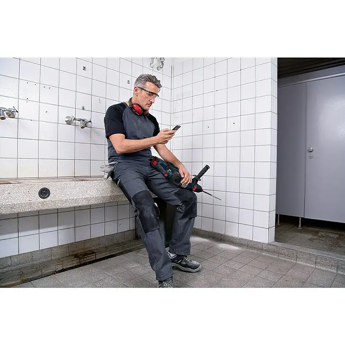 Bosch Professional AMPShare 18V Akku-Bohrhammer GBH 18V-34 CF - 18 V, 2 Akkus, 5,5 Ah, 5,8 J - Arbeiter hält einen blau-schwarzen Bohrhammer mit großem Bohrer, blickt auf sein Telefon in gefliestem Technikraum.