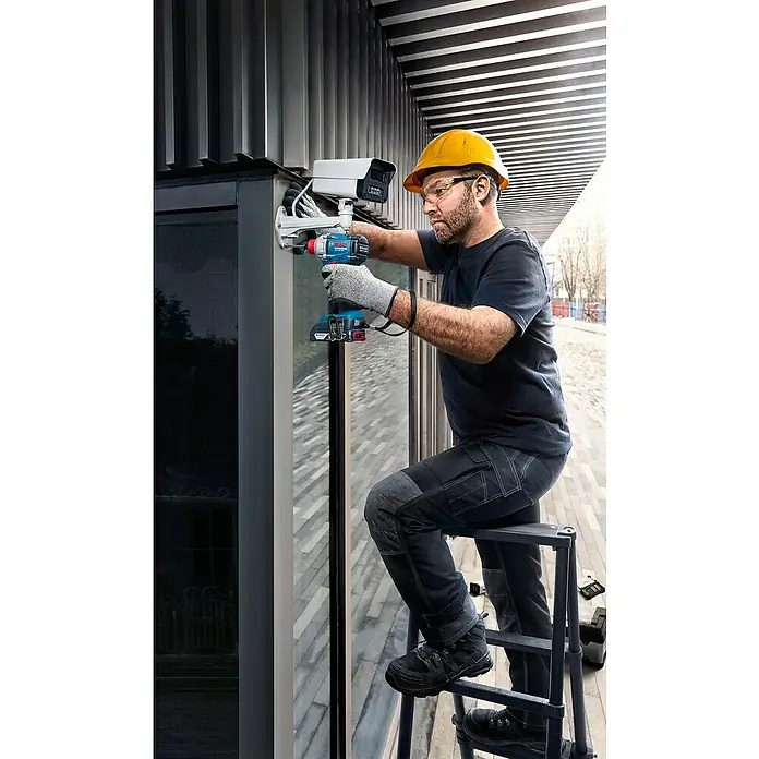 Bosch Professional AMPShare 18V Akku-Drehschlagschrauber GDX 18V-285 C - 18 V, Ohne Akku, 285 Nm - Mann mit Bauhelm montiert mit einem blau-schwarzen Bosch Akku-Bohrschrauber eine weiße Überwachungskamera an eine Fassade von einer Leiter aus.