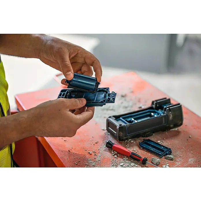 Bosch Professional USB-Akku-Adapter PRO BA 3.7V 1.0Ah A - Akkuspannung: 3,7 V, Höchste Leistung Einzelakku: 5 Wh - Hände montieren ein dunkelblaues und schwarzes Kunststoffgehäuse, möglicherweise für Elektronik, auf einer staubigen Oberfläche.