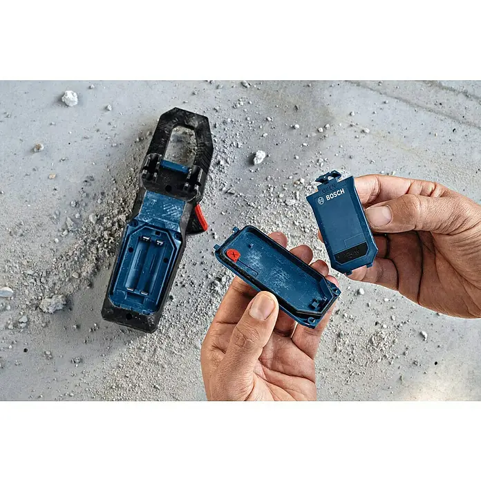 Bosch Professional USB-Akku-Adapter PRO BA 3.7V 1.0Ah A - Akkuspannung: 3,7 V, Höchste Leistung Einzelakku: 5 Wh - Hände halten einen blauen Bosch-Akkupack und dessen Abdeckung, neben einem geöffneten Bosch-Zangenmessgerät auf staubigem Betonboden.