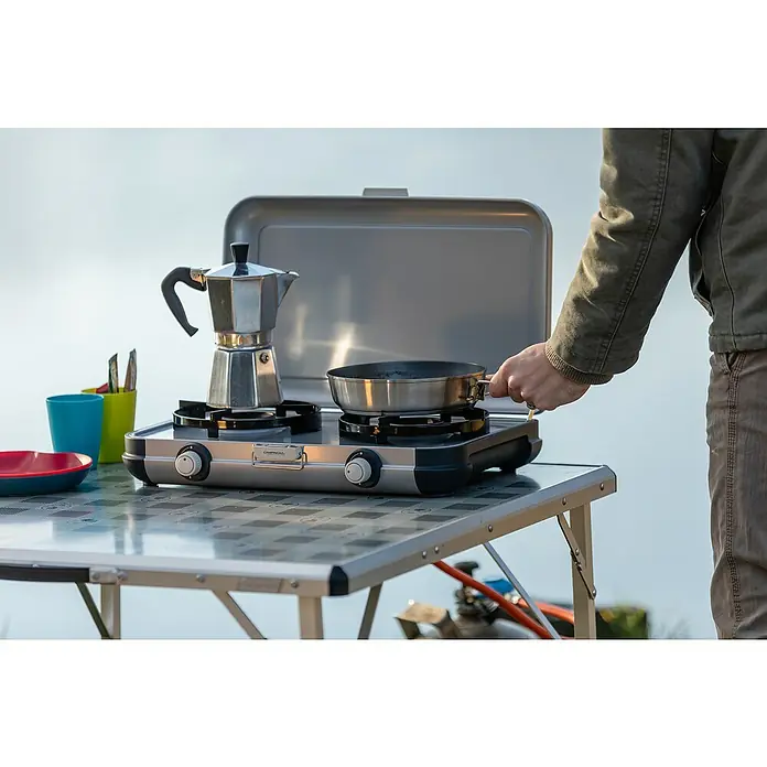 Campingaz Gaskochfeld Kitchen 2 - 2 Brenner, 2.000 W, L x B x H: 35 x 49 x 10 cm - Person kocht auf einem silbernen Campingaz-Doppelbrenner-Gaskocher, hält eine Pfanne. Eine Mokkakanne steht ebenfalls auf dem Kocher.