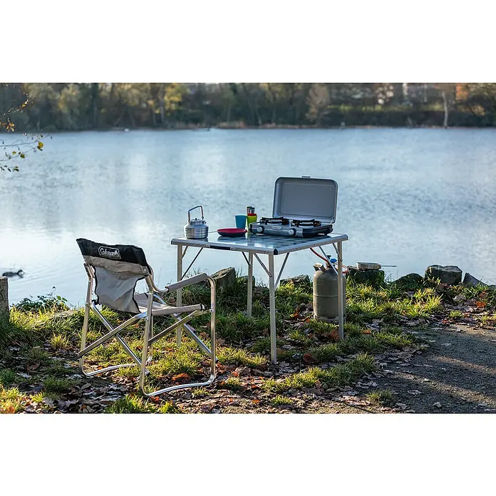 Campingaz Gaskochfeld Kitchen 2 - 2 Brenner, 2.000 W, L x B x H: 35 x 49 x 10 cm - Ein tragbarer Campingtisch mit Gaskocher, Kessel, Tassen und Tellern, neben einem Campingstuhl am See, bereit zum Kochen im Freien.