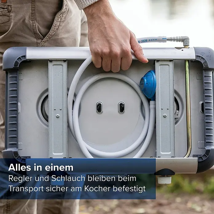 Campingaz Gaskochfeld Kitchen 2 - 2 Brenner, 2.000 W, L x B x H: 35 x 49 x 10 cm - Eine Person trägt einen geschlossenen, kompakten silber/grauen tragbaren Gaskocher, Schlauch und Regler sind sicher verstaut.