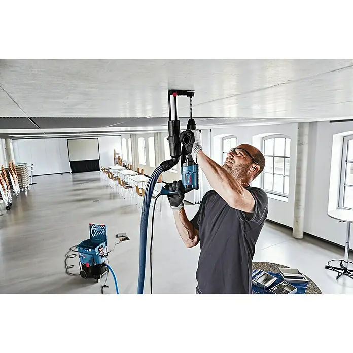 Bosch Professional Absaugvorrichtung GDE 16 Plus - Passend für: Bosch Professional Bohrhämmer 2 & 3 kg - Ein Mann bohrt mit einer blauen Bosch Bohrvorrichtung Löcher in eine Betondecke in einem leeren Raum.