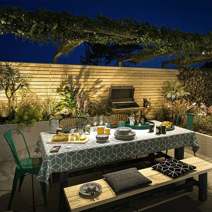 Philips Aplique exterior Bellini - 25 W, L x An x Al: 16,5 x 21,5 x 28 cm, IP44 - Un área de comedor exterior de noche con una mesa puesta, con un mantel azul geométrico, sillas verdes y un banco de madera con cojines.