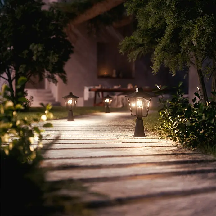 Philips Sobremuro exterior Kinno - 25 W, Color del marco: Negro, 17 x 17 x 39 cm, Sin bombillas - Luces negras de jardín iluminan un camino de madera por la noche, creando un ambiente exterior cálido y acogedor.