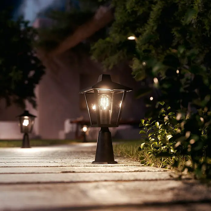 Philips Sobremuro exterior Kinno - 25 W, Color del marco: Negro, 17 x 17 x 39 cm, Sin bombillas - Farol de jardín negro con bombilla de filamento iluminando un camino de piedra por la noche.