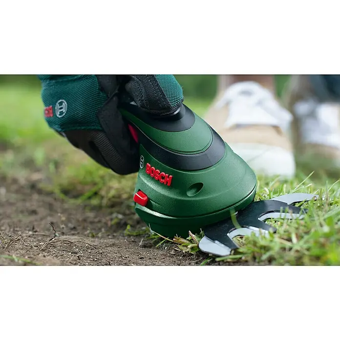 Bosch Akku-Gras- & Strauchschere Isio 3 - 3,6 V, 1 Akku, Schwertbreite: 8 cm - Behandschuhte Hand hält grüne und schwarze Bosch Grasschere, schneidet Gras und Erde am Boden.