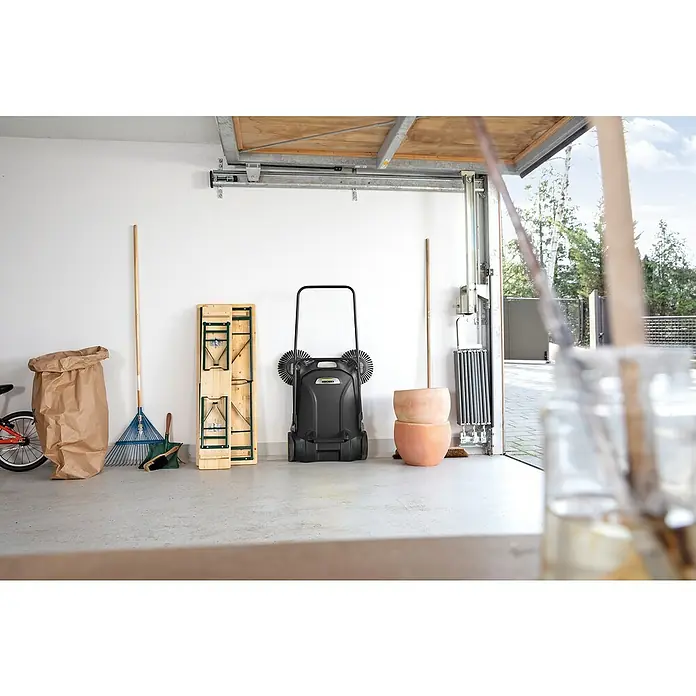 Kärcher Handkehrmaschine S 4 Twin Go!Further - Flächenleistung: 2.400 m²/h, 20 l - Ein schwarzer Kärcher Kehrbesen mit gelbem Logo steht in einer Garage, umgeben von Gartengeräten.
