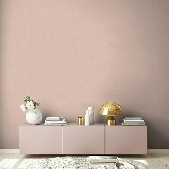 A.S. Création New Walls Vliestapete Textil - Rosé, Uni, 10,05 x 0,53 m - Eine stilvolle Inneneinrichtung mit einer rosa Anrichte, einer goldenen Lampe und dekorativen Vasen und Büchern.