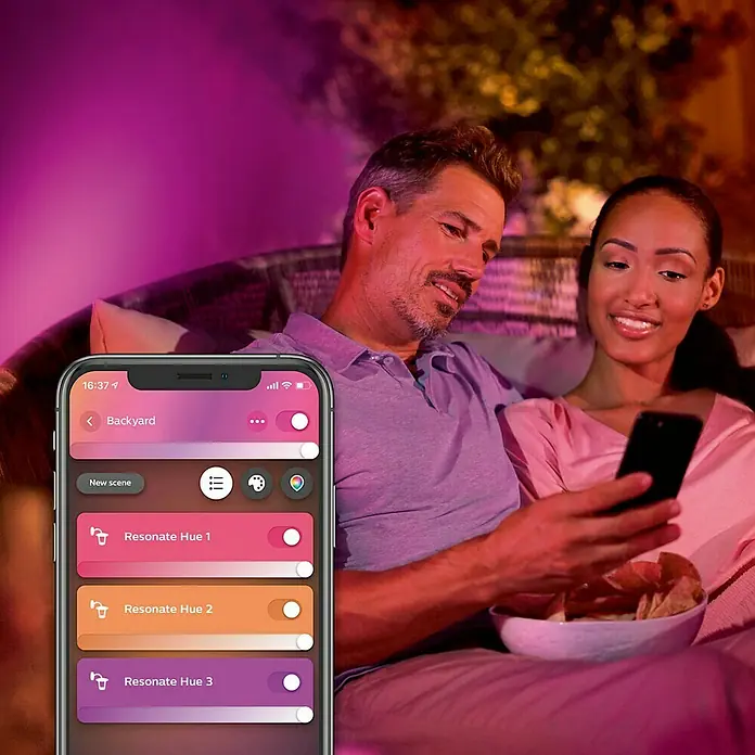 Philips Hue LED-Außenwandleuchte White & Color Ambiance Resonate Up & Down - 16 W, 9,3 x 12 x 18 cm, Schwarz, IP44 - Ein Paar entspannt sich im Hinterhof mit einem Smartphone, das die Philips Hue Lichtsteuerung zeigt.