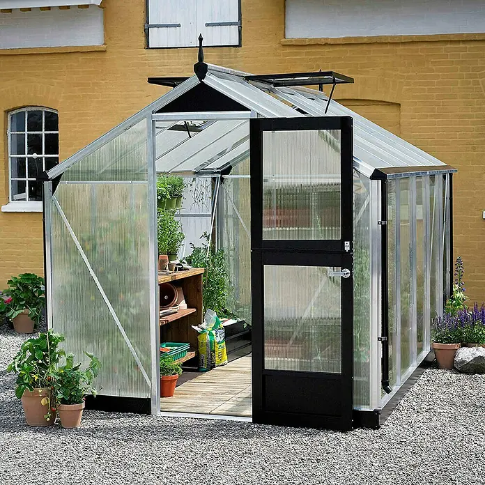 Juliana Gewächshaus Compact - Außenmaß inkl. Dachüberstand (B x T): 224 x 295 cm, Material Seitenwand: Kunststoff, 10 mm, Silber/Schwarz - Ein geräumiges Gewächshaus mit geöffneter Tür, umgeben von Pflanzen und Kies, lädt zum Gärtnern ein. Ideal für den Anbau von Gemüse und Blumen.