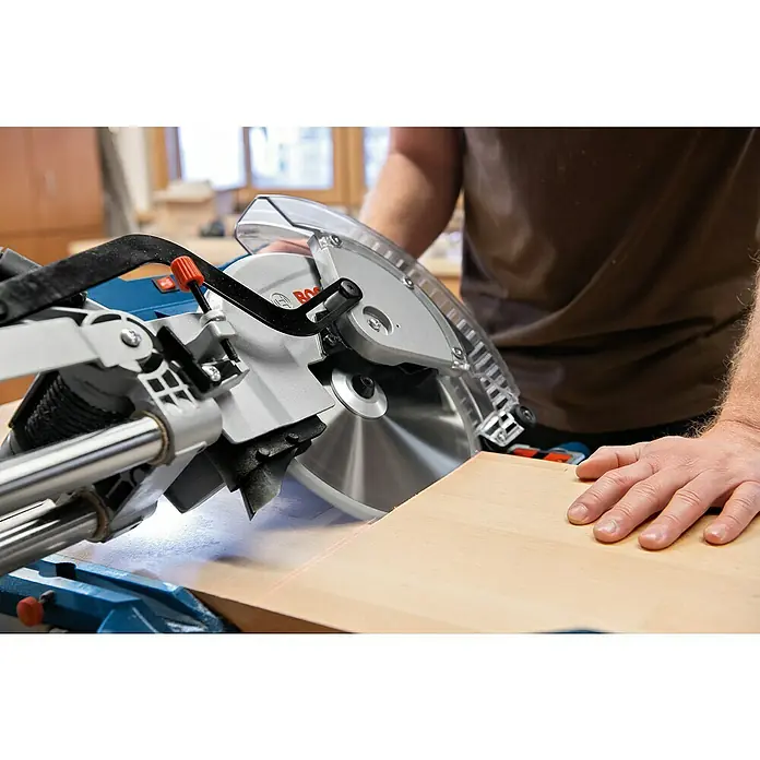 Bosch Professional Kapp- & Gehrungssäge GCM 305-254 D - 1.500 W, Durchmesser Sägeblatt: 254 mm, Leerlaufdrehzahl: 4.500 U/min - Blau-silberne Kappsäge mit transparentem Schutzblatt schneidet Holz, geführt von einer roten Laserlinie. Eine Hand hält das Holz.