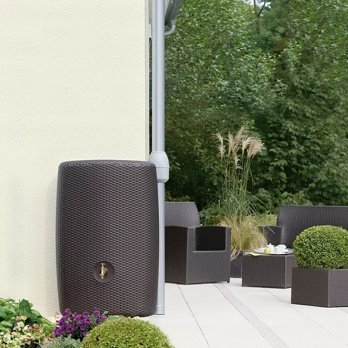 Garantia Exklusive Line Regenspeicher Sunda - 300 l, Mocca - Eine elegante, dunkle Regenwassertonne im Rattan-Design mit Messinghahn steht neben einem Fallrohr an einer hellen Hauswand.