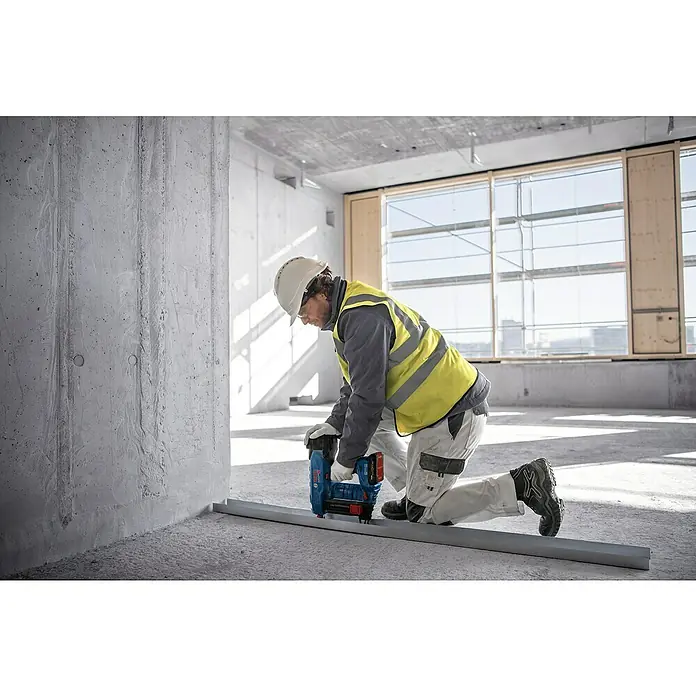 Bosch Professional Akku-Nagler GNB 18V-38PRO - 18 V, 2 Akkus, Länge Nagel: 13 mm - 38 mm - Ein Arbeiter kniet auf Betonboden und verwendet einen blauen und schwarzen Bosch Akku-Nagler entlang einer Metallschiene.