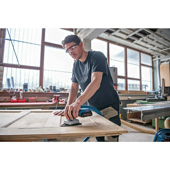 Bosch Professional AMPShare 18V Akku-Schwingschleifer GSS 18V-13 - Anzahl Akkus: Ohne Akku, Leerlaufschwingzahl: 12.000 U/min - 20.000 U/min, 18 V - Mann schleift ein Holzbrett mit einem blauen und schwarzen Bosch Akku-Exzenterschleifer, verbunden mit einem Absaugschlauch.