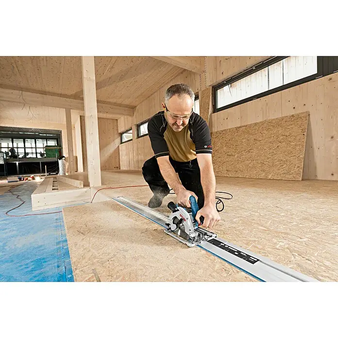 Bosch Professional Handkreissäge GKS 55+ GCE - 1.350 W, Durchmesser Sägeblatt: 165 mm - Mann kniet, sägt OSB-Platte mit blauer und silberner Tauchsäge auf Führungsschiene in Holzgebäude.