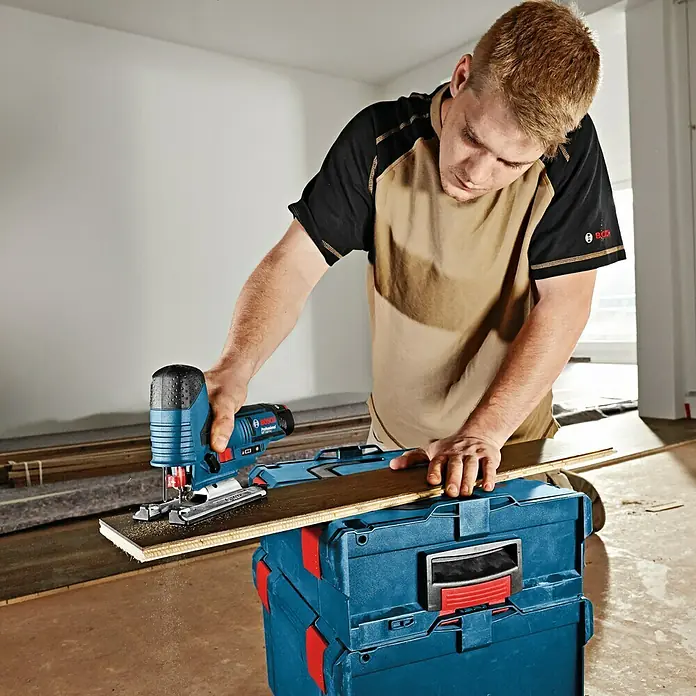 Bosch Professional Akku-Stichsäge GST 12V-70 - 12 V, Li-Ionen, Ohne Akku, Leerlaufhubzahl: 1.500 /min - 2.800 /min - Ein Mann benutzt eine Bosch Stichsäge, um ein Stück Holz über einer blauen Werkzeugkiste zu schneiden.