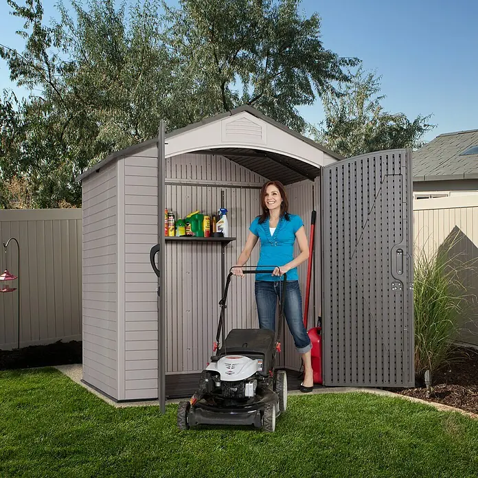 Lifetime Gerätehaus Starlet - 215 cm, 140 cm, Kunststoff, Anthrazit/Grau - Eine Frau schiebt einen Rasenmäher in einen grauen Gartenschuppen mit offenstehender Tür, der Platz und Aufbewahrungsmöglichkeiten bietet.