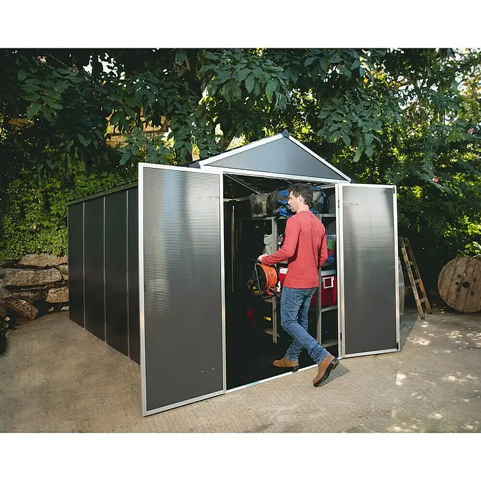 Palram – Canopia Gartenhaus Rubicon - Außenmaß inkl. Dachüberstand (B x T): 1,85 x 3,78 m, Kunststoff, Grau - Mann betritt einen dunkelgrauen Gartenlager, der mit Regalen und offenen Türen ausgestattet ist.