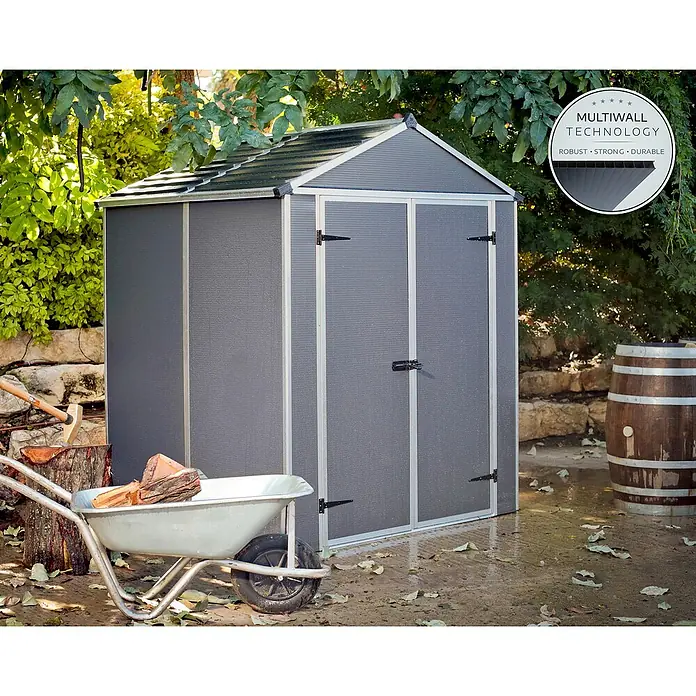 Palram – Canopia Gartenhaus Rubicon - Außenmaß inkl. Dachüberstand (B x T): 1,85 x 1,54 m, Kunststoff, Grau - Ein dunkelgraues Gartenhaus mit hellgrauem Rahmen und Dach in einem Garten, neben einer Schubkarre mit Holz.