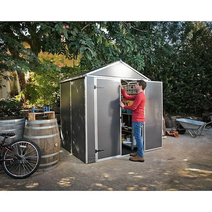 Palram – Canopia Gartenhaus Rubicon - Außenmaß inkl. Dachüberstand (B x T): 1,85 x 1,54 m, Kunststoff, Grau - Ein Mann öffnet einen grauen Garten-Schuppen mit geriffelten Platten und einem silbernen Rahmen zur Gartenlagerung.