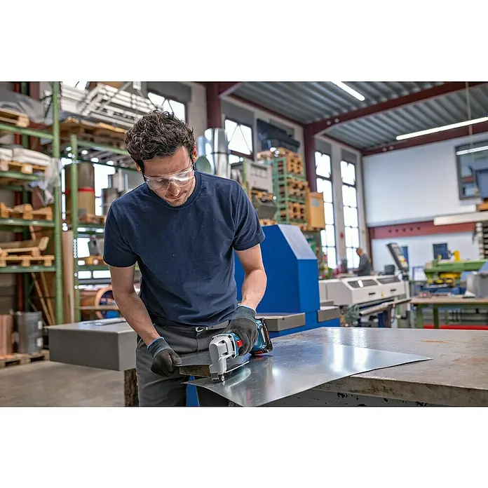 Bosch Professional AMPShare 18V Akku-Blechschere Heavy Duty GSC 18V-16 E - 18 V, Ohne Akku, Leerlaufhubzahl: 3.200 /min - Mann mit Schutzbrille und Handschuhen benutzt eine blaue und schwarze Akku-Blechschere zum Schneiden eines silbernen Blechs in einer Werkstatt.