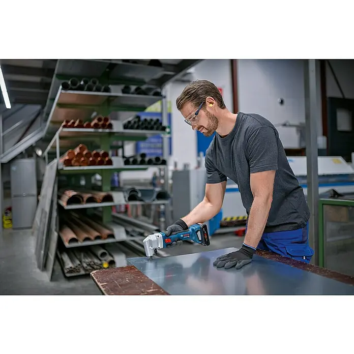 Bosch Professional Akku-Nager GNA 18V-16 E PRO - 18 V, 2 Akkus, 12 Ah - Ein Mann schneidet mit einer blauen und schwarzen Bosch Akku-Blechschere präzise ein silbernes Metallblech auf einem Holztisch.
