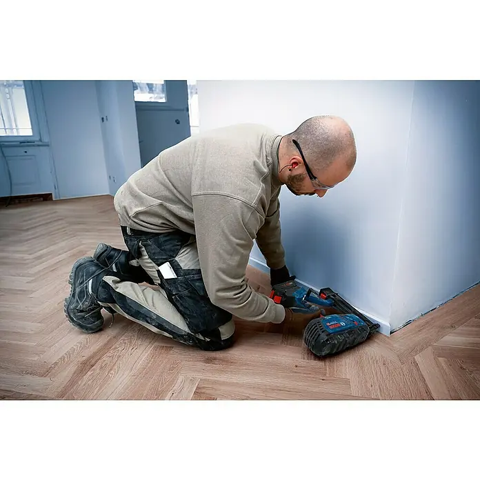 Bosch Professional Akku-Nagler GNH 18V-50 M PRO SOLO - 18 V, Ohne Akku, Länge Nagel: 16 mm - 50 mm - Eine Person kniet und verwendet ein blau-schwarzes Bosch Nagler zur Installation von weißer Sockelleiste.