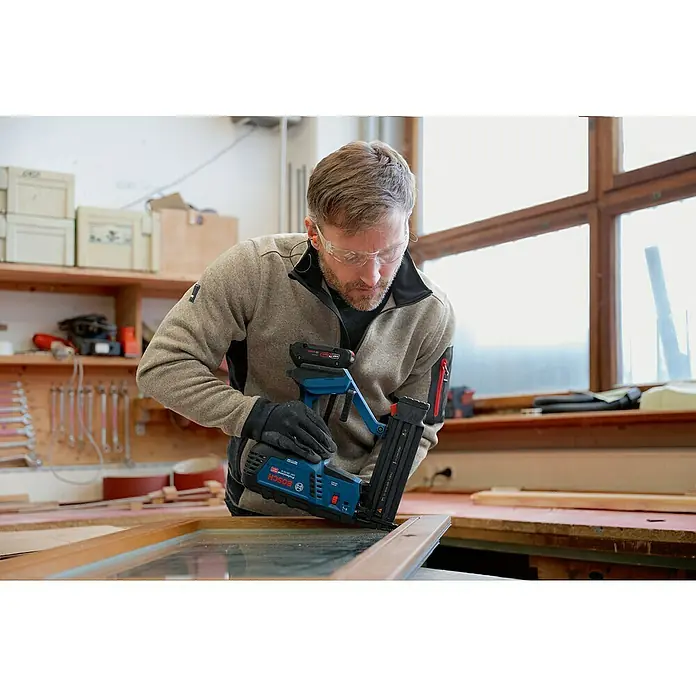 Bosch Professional Akku-Nagler GNH 18V-50 M PRO SOLO - 18 V, Ohne Akku, Länge Nagel: 16 mm - 50 mm - Mann nutzt blauschwarzen Bosch Professional Akku-Nagler an Holzfensterrahmen in einer Werkstatt.