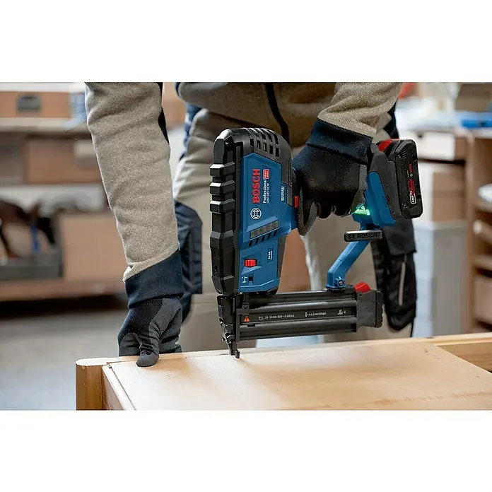 Bosch Professional Akku-Nagler GNH 18V-50 M PRO SOLO - 18 V, Ohne Akku, Länge Nagel: 16 mm - 50 mm - Behandschuhte Hände halten ein blau-schwarzes Bosch Akku-Nagelgerät, dessen Spitze auf eine helle Holzoberfläche gedrückt wird.