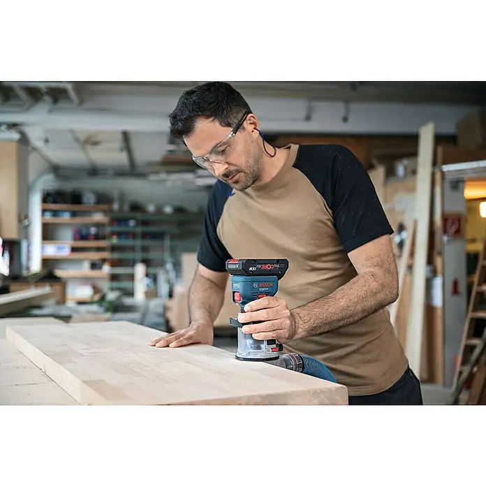 Bosch Professional AMPShare 18V Akku-Kantenfräser GKF 18V-8 PRO SOLO - 18 V, Ohne Akku, Leerlaufdrehzahl: 10.000 U/min - 30.000 U/min - Mann verwendet eine blau-schwarze Bosch Professional Akku-Oberfräse auf einem Holzbrett.