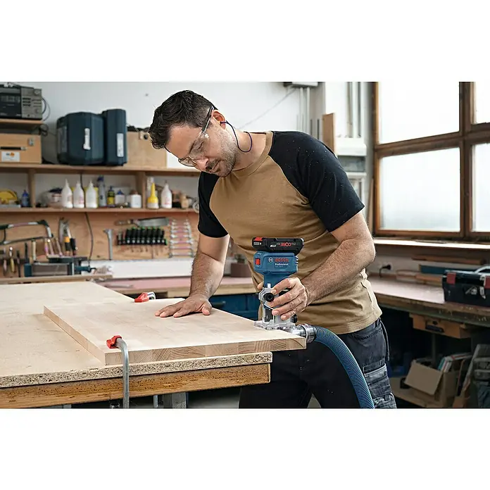 Bosch Professional AMPShare 18V Akku-Kantenfräser GLF 18V-8 PRO SOLO - 18 V, Ohne Akku, 10.000 U/min - 30.000 U/min, Inkl. L-Boxx - Mann mit Schutzbrille nutzt blaue und schwarze Bosch Professional Akku-Oberfräse mit Staubabsaugung an Holzbrett.