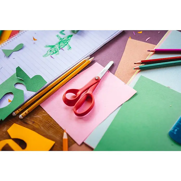 Fiskars Bastelschere Classic - Rot - Kinderbastelbedarf: rote Schere, Buntstifte und Papier auf einem Holztisch, dazu ein Notizbuch mit Krokodilzeichnung.