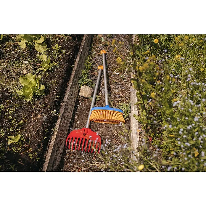 Fiskars Kinder-Besen Aufräum-Set - Länge: 863 mm, 2 -tlg. - Rote Kunststoffharke und blau-gelber Besen mit silbernen Stielen liegen auf einem Gartenweg neben grünen Pflanzen.