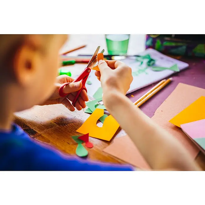 Fiskars Bastelschere Classic - Rot - Nahaufnahme von Kinderhänden, die mit roter Schere oranges Papier bei einer Bastelaktivität schneiden.