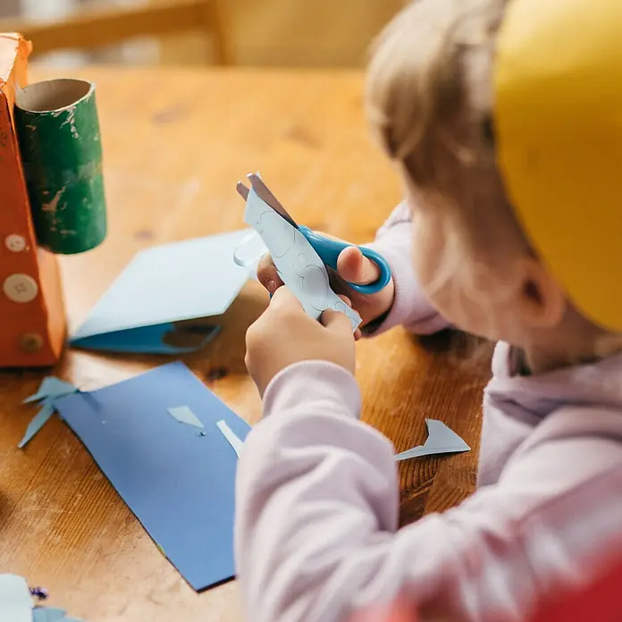 Fiskars Bastelschere - Länge: 131 mm, Türkis - Kinderhände schneiden mit einer blauen Schere Papier an einem Holztisch, umgeben von Bastelmaterialien.