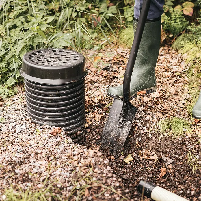Fiskars Ergonomic Gärtnerspaten Pro - Arbeitsbreite: 15,8 cm, Länge: 1.307 mm - Schwarzer, gerippter Kunststoff-Drainageschacht mit geschlitztem Deckel wird im Freien neben einer Schaufel und einer Person in Stiefeln installiert.