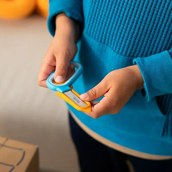 Fiskars Bastel-Set Kreativset für Kinder aus Pappe - 2 -tlg. - Kinderhände halten einen blauen und gelben Fiskars Cardboard Creator Sicherheits-Cutter.
