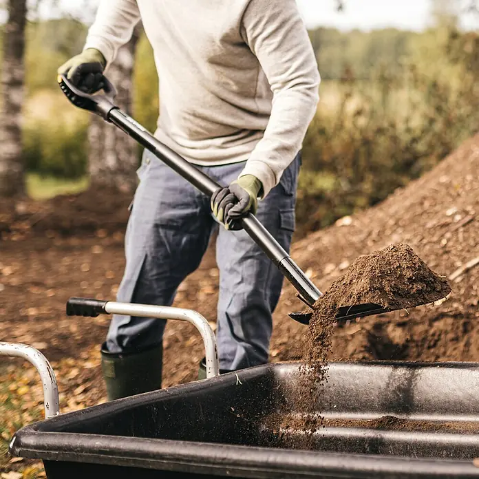 Fiskars Ergonomic Erdschaufel Pro - Material Gerätekopf: Stahl, Länge: 1.317 mm - Person mit Handschuhen schaufelt braune Erde mit einer schwarzen Schaufel in eine schwarze Schubkarre.