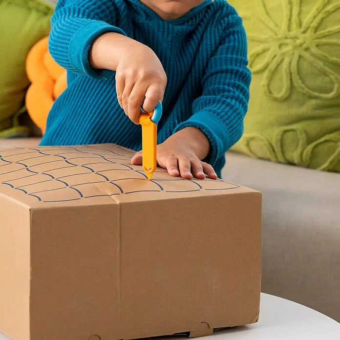 Fiskars Bastel-Set Kreativset für Kinder aus Pappe - 2 -tlg. - Ein Kind schneidet mit einem orange-blauen Sicherheitsmesser ein gezeichnetes Muster auf einen braunen Karton.