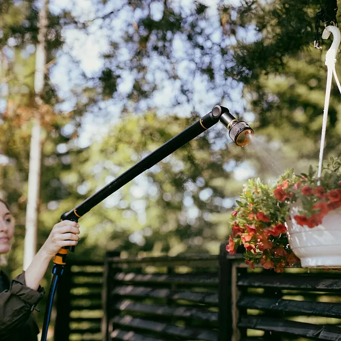 Fiskars Gießstab Comfort - Länge: 76 cm, Aluminium - Eine Person bewässert rote Hängeblumen mit einer schwarzen und orangefarbenen langen Gießlanze, die Wasser sprüht.