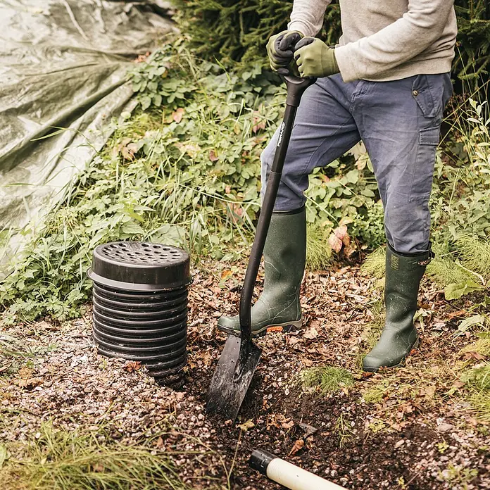 Fiskars Ergonomic Gärtnerspaten Pro - Arbeitsbreite: 15,8 cm, Länge: 1.307 mm - Schwarzer Wellrohr-Drainageschacht mit perforiertem Deckel wird draußen neben einer Schaufel installiert.