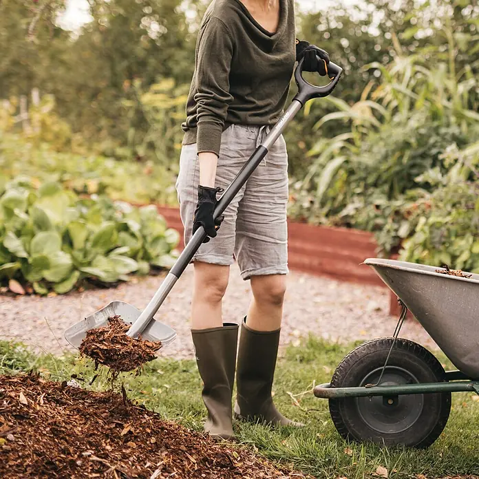Fiskars Ergonomic Schaufel - Breite: 230 mm, D-Griff - Person in Gartenstiefeln und Handschuhen schaufelt Holzspäne mit einer grau-schwarzen Fiskars Schaufel in eine Schubkarre.