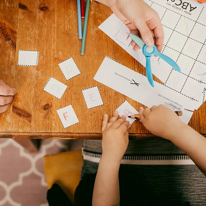 Fiskars Bastel-Set Anfängerschere & Bastelheft - 2 -tlg. - Erwachsenen- und Kinderhände schneiden und malen Alphabetbuchstaben mit Kinderschere und Stiften auf einem Holztisch.