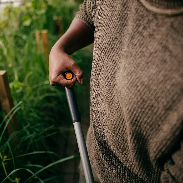 Fiskars Jäter Ergonomic - L x B x H: 1.660 x 230 x 260 mm - Eine Hand hält ein Gartengerät mit orange-schwarzem Griff, Person trägt strukturiertes Hemd.