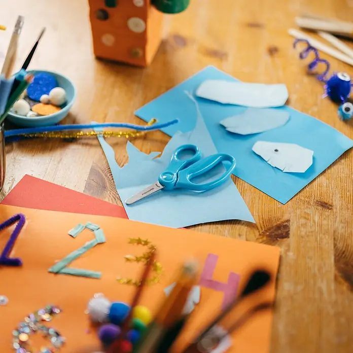Fiskars Bastelschere - Länge: 131 mm, Türkis - Bastelmaterialien auf einem Holztisch: blaues Papier, Schere, Pfeifenreiniger und verzierte Zahlen.