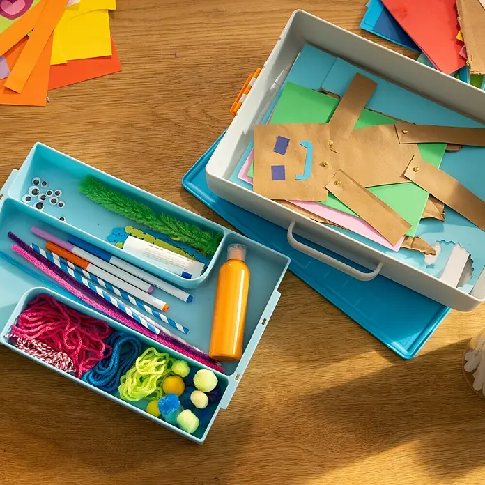 Fiskars Werkzeugkoffer Bastelbox Kids - L x B x H: 386 x 77 x 252 mm, Kunststoff - Buntes Bastelzubehör wie Garn, Wackelaugen und Marker in blauen und weißen Aufbewahrungsboxen auf einem Holztisch.