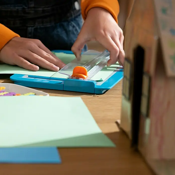 Fiskars Schneidegerät Kids Junior Papiertrimmer 16L - L x B x H: 289 x 35 x 177 mm - Eine Person benutzt einen blauen Papierschneider mit orangefarbenem Schieber, um hellgrünes Papier zu schneiden.