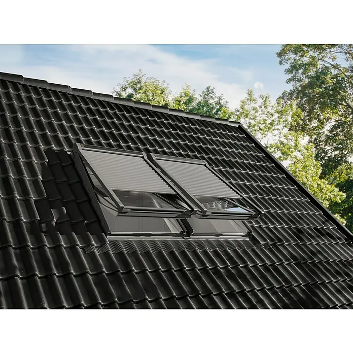 Velux Dachfenster-Rollladen Elektro SML CK06 0000K - Passend für Fenstergröße: CK06, C06, Passend für Fenstertyp: GGL, GGU, GPL, GPU, GHL, GHU, GZL, GLL, GLU, VU und VKU - Zwei graue Außenrollläden auf Dachfenstern in einem dunklen Ziegeldach, teilweise geöffnet, mit Himmel und Bäumen im Hintergrund.